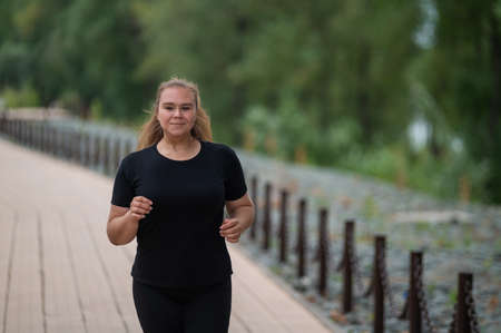 An obese young woman jogging outdoors. Fat beautiful smiling girl in a black tracksuit is engaged in fitness for weight loss on the waterfront. A woman runs on a summer day.の写真素材