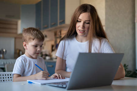 Caucasian woman helps boy learn lessons. The tutor checks the childs homework. Individual home-based quarantine. A diligent boy writes a dictation under the supervision of a teacher.の写真素材