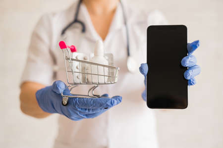 A faceless female doctor in a medical coat with a stethoscope holds a mini shopping trolley full of different medicines and a smartphone with a blank screen. Mobile application for the purchase of medicines. Online pharmacyの写真素材