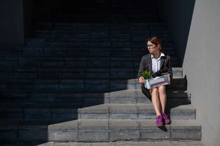 Depressed woman dressed in medical mask is fired and is sitting on the stairs with a box of personal belongings. Female office worker in suit and sneakers outdoors. Unemployment in the economic crisisの写真素材