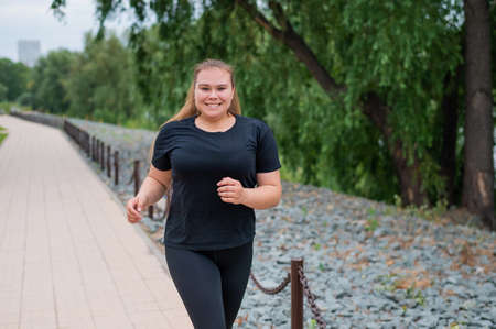 An obese young woman jogging outdoors. Fat beautiful smiling girl in a black tracksuit is engaged in...
