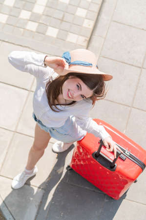 Top view on beautiful young caucasian woman in summer hat posing with red suitcase outdoors. Happy...