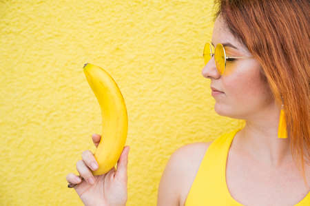 Portrait of caucasian redhead woman in sunglasses holding a banana in her hands. Beautiful seductive girl posing on a yellow background.の写真素材