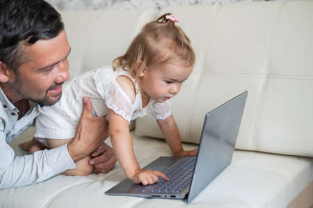 Little girl and caring father are watching cartoons on a laptop while lying on the couch at home. A...