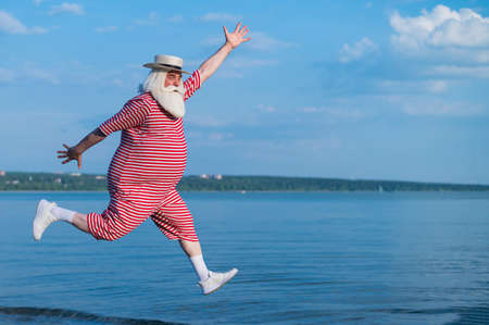 An elderly man in a striped retro swimsuit runs and jumps along the beach. A cheerful gray-haired...