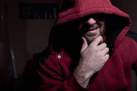 Portrait of an evil bearded man in a hoodie in the dark. Male criminal hides his eyes under the...