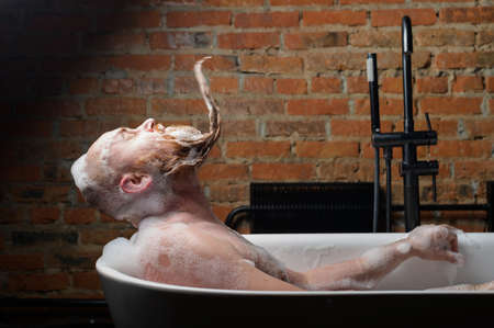 Close-up portrait of funny red-bearded bald man takes a bath with foam on the background of a brick...
