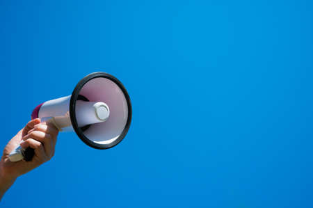 Megaphone in a female hand on a blue background. No people. Cropped photo. A woman holds a sound...