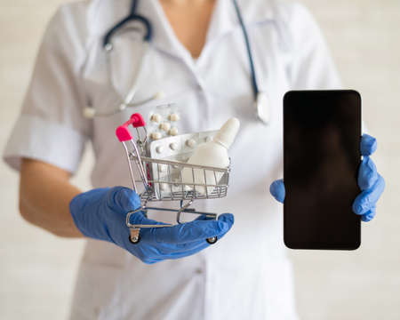 A faceless female doctor in a medical coat with a stethoscope holds a mini shopping trolley full of different medicines and a smartphone with a blank screen. Mobile application for the purchase of medicines. Online pharmacyの写真素材