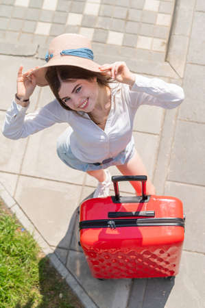 Top view on beautiful young caucasian woman in summer hat posing with red suitcase outdoors. Happy smiling girl goes on vacation with a large luggage bag. Charming lady in shorts on a hot sunny day.の写真素材