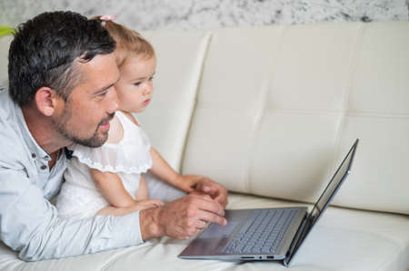Little girl and caring father are watching cartoons on a laptop while lying on the couch at home. A man shows his little daughter photographs on a computer. Beloved daddy.の写真素材