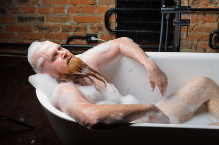 Close-up portrait of funny red-bearded bald man takes a bath with foam on the background of a brick...