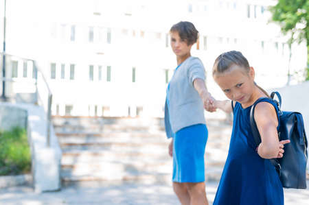 A little unhappy girl with a backpack pulls her mother by the hand does not want to go to school. A woman leads a resisting daughter to school. The conflict of generations.の写真素材