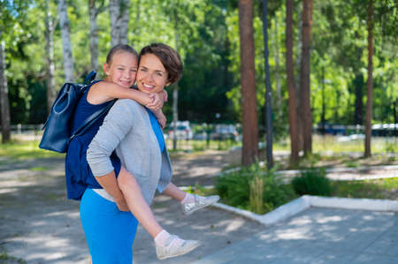 A beautiful caucasian woman stands outdoors and holds daughter on her back. A cheerful smiling girl sits on her mother back while walking in the park. Friendly loving family.の写真素材