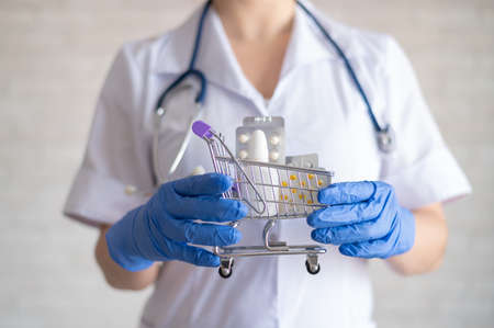 A faceless female doctor in rubber gloves holds a mini shopping trolley full of different medicines. The concept of buying drugs in a pharmacy.の写真素材