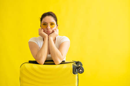Unhappy woman in sunglasses hugs a suitcase on a yellow background. An upset girl missed her flight on a plane. Cancellation of all flights due to coronavirus. Lack of summer vacation.の写真素材