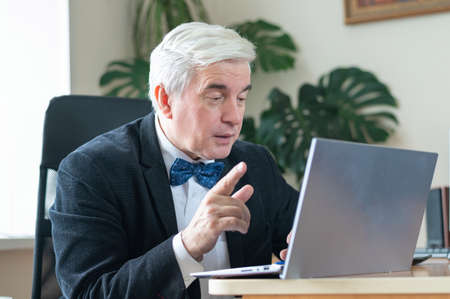 Attractive mature man in a suit and a bow tie communicates on a laptop webcam. Confident gray-haired male boss is hosting an online conference. Business man is conducting remote negotiations.の写真素材