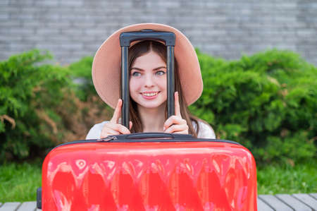 Young beautiful caucasian woman in a hat looks out of a red suitcase with a handle. Happy girl goes on a trip with luggage. Summer vacation concept.の写真素材