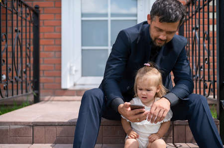 Caucasian man in a business suit looks at the mobile screen with daughter on the stairs. A little girl with a loving father watching cartoons on a smartphone while sitting on the porch of the house.の写真素材