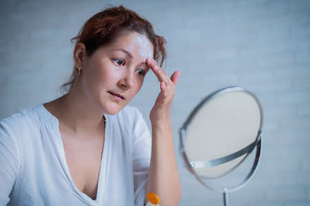 Portrait of a Caucasian woman with vitiligo uses sunscreen. A girl with a white pigment spot on her forehead looks in the mirror and is smeared with cream.の写真素材