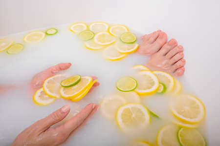 Faceless beautiful young woman takes a bath with milk lemons and lime. Cropped photo. Close-up of female hands and feet in a skin whitening bath.の写真素材