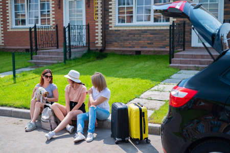 Three Caucasian women and a dog are going on a road trip. The girls are loading their suitcases into...