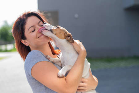 Happy red-haired woman hugs her little dog for a walk outdoors. Jack Russell Terrier licks the face of the female owner.の写真素材