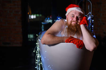 Humorous image of an evil santa claus taking a bath with christmas lights. A man with a red beard in a Santa Claus hat and underwear is holding a garland in his hands.の写真素材