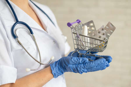 A faceless doctor with a medical coat and a stethoscope holds a small shopping cart full of medicines. Pharmacy concept.の写真素材