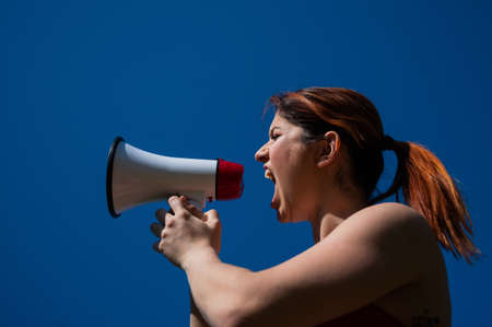Red-haired Caucasian woman yelling into a megaphone against a blue sky background. The girl makes an...