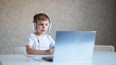 Cute little boy is watching cartoons or a movie on a laptop. Caucasian child sits at a table at home...