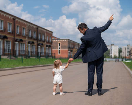 Rear view of a man in a black suit walks with his little daughter outdoors. A little girl in a white dress holds her fathers hand. Paternal loveの写真素材