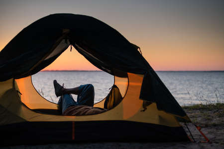 A woman lies in a tourist tent and enjoys the sunset by the river.
