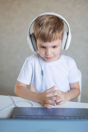 A diligent boy in headphones sits at a desk and watches an online lesson on a laptop.の写真素材
