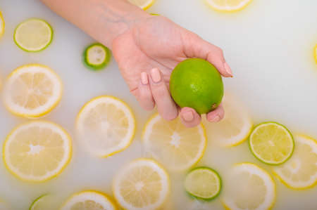 Close-up of a female hand holding a lime over white water with lemon slicesの写真素材