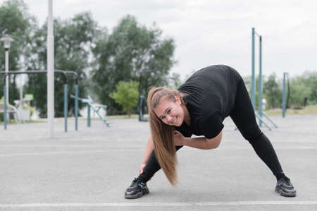 Fat young woman doing stretching exercises outdoors. Overweight girl makes bends before jogging.