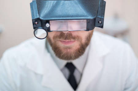 Male bearded doctor in a medical coat and with a magnifying head strap w lights sitting on the desk. Optometrist equipment. Medical office.の写真素材