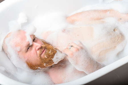Close-up portrait of funny red-bearded bald man taking a bath with foam. Cheerful naughty guy at spa treatments. A parody of glamorous girls.の写真素材