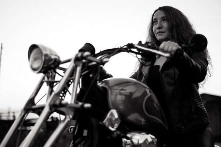 Curly red-haired woman in a black leather jacket sits on a motorcycle. Portrait of a serious girl driving a bike.の写真素材