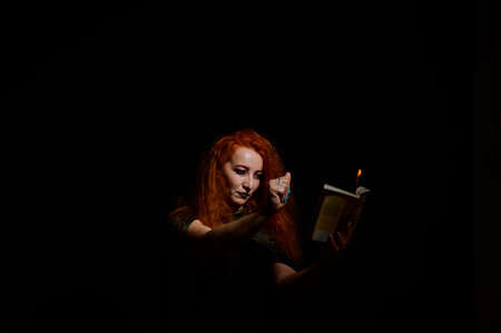 An ominous witch sets her spell book on fire with the power of thought. Red-haired woman conjures for Halloween. Flames on the hands.の写真素材