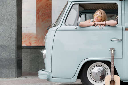 A beautiful young hippie woman in yellow sunglasses looks out the window of a blue vintage minivan