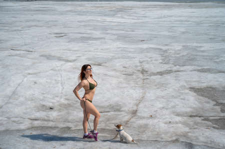 A red-haired woman in a bikini walks with a dog on a snowy beachの写真素材