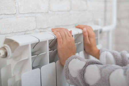 A woman wearing in pajamas warms cold hands on the heating radiatorの写真素材