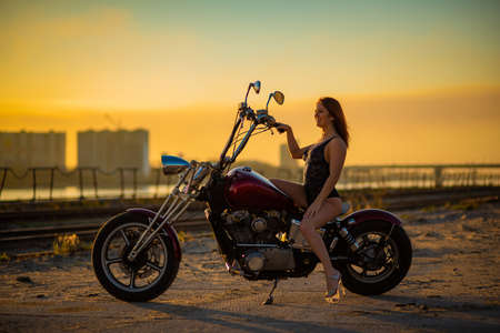 Red-haired woman in sexy lingerie in high heels sits on a motorcycle. Attractive red-haired girl sits on a motorcycle at sunsetの写真素材