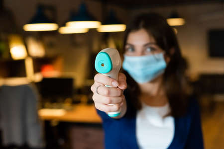 A female office worker in a mask holds a contactless electronic thermometer
