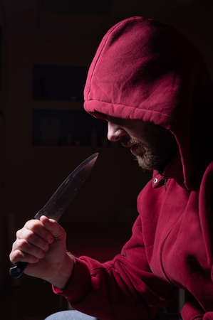 A dangerous bearded man in a red hood holds a sharp knife near his face. A criminal or a killer with knives in the dark.の写真素材