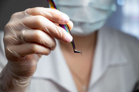 Lashmaker in a mask holds a bunch of eyelashes with tweezers. Close-up of a working tool of a master...