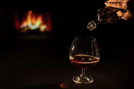 Woman pours cognac into a glass on the background of the fireplace.の写真素材