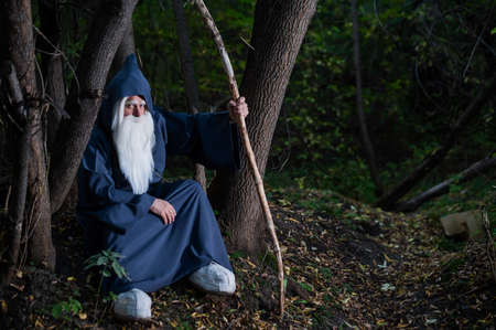 A wizard with a long gray beard and a cloak in a deep forest. An elderly man in a witcher costume