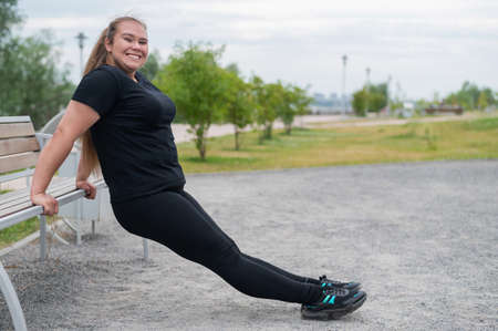Young fat woman doing push-ups from the bench outdoors. Chubby girl doing fitness exercises.